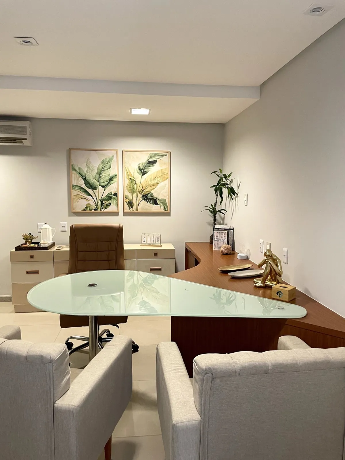 Consultório da Clínica Proteger Saúde, com poltronas claras, mesa de vidro e quadros de folhagem